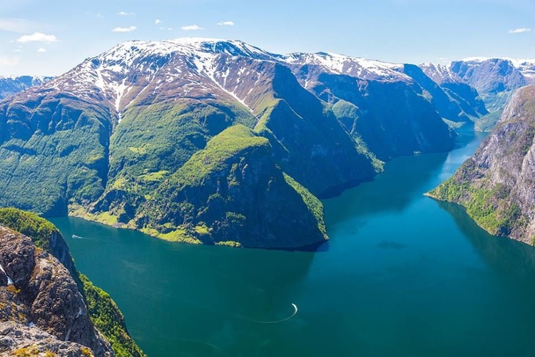 Aerial view of a deep blue fjord surrounded by steep, rugged mountains with patches of snow and lush green slopes under a clear sky