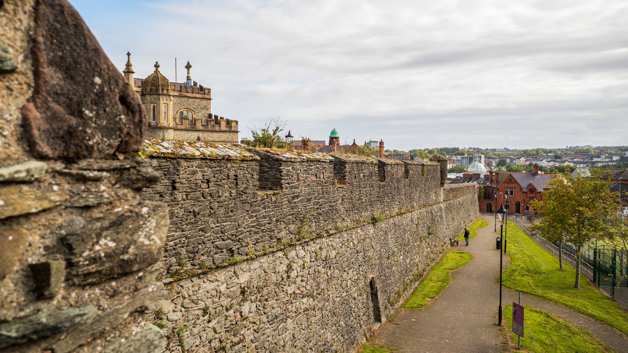 View of Derry’s historic city walls with stone ramparts, greenery, and city buildings under a cloudy sky