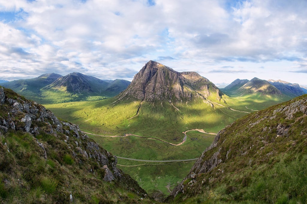 Scenic landscape of the Scottish Highlands featuring rolling green mountains, a calm loch, and dramatic cloudy skies.