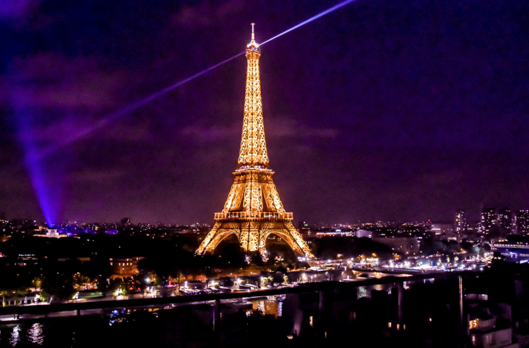 The Eiffel Tower glowing with golden lights at night against a deep purple sky, with a bright spotlight beaming from the top and the Paris cityscape illuminated below.