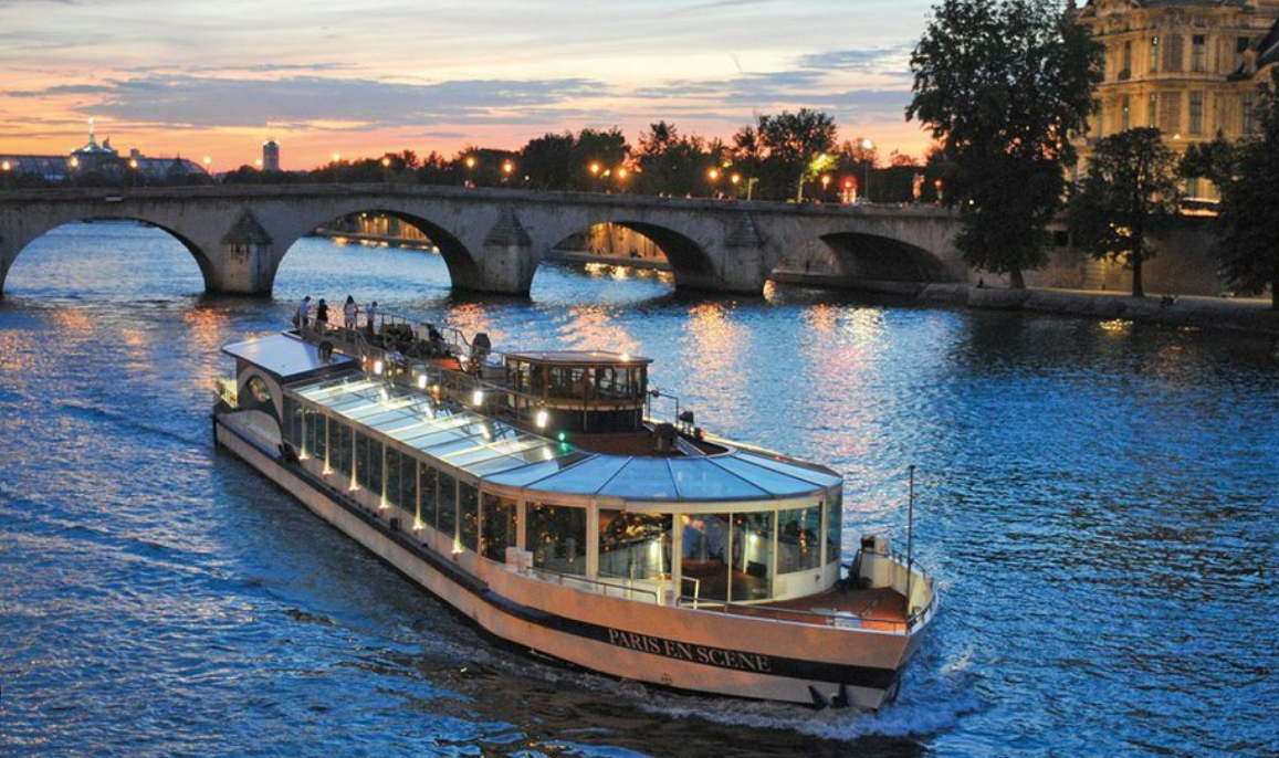 A glass‑roofed sightseeing boat cruising along the Seine River in Paris at sunset, with warm lights reflecting on the water and a stone arch bridge in the background.