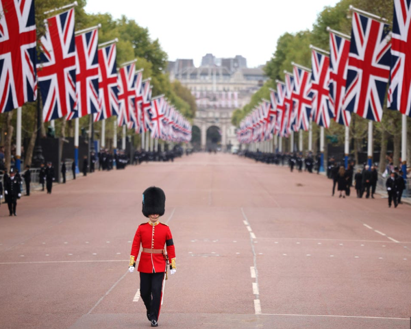 Beefeater in traditional red and gold uniform walking down a London street towards Buckingham Palace, royal guard heading for duty with historic palace and city traffic in the background.