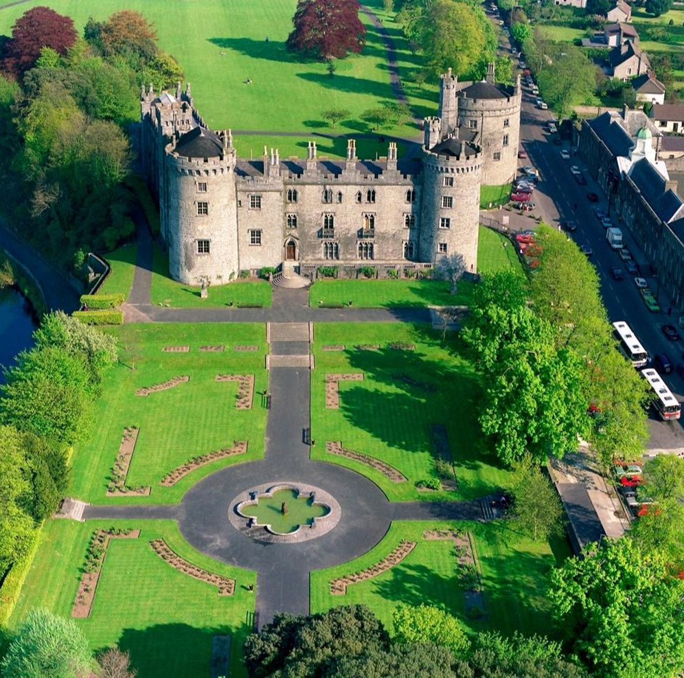 Kilkenny Castle in Ireland with its grand stone façade, towers, and surrounding green lawn under a bright sky.