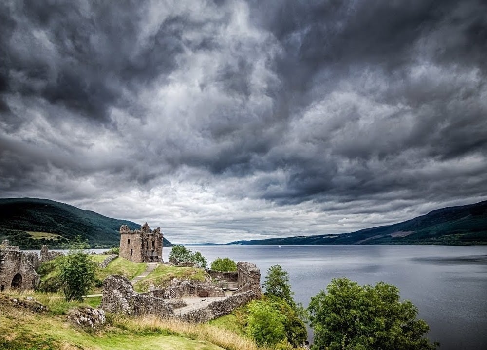 Scenic view of Loch Ness in Scotland with calm blue water surrounded by rolling green hills and a cloudy sky.
