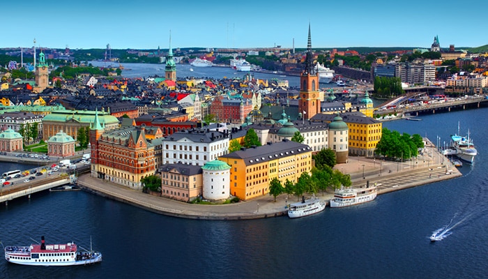 Colorful historic waterfront buildings on a small peninsula surrounded by boats and deep blue water, with a wide cityscape and church towers in the background.