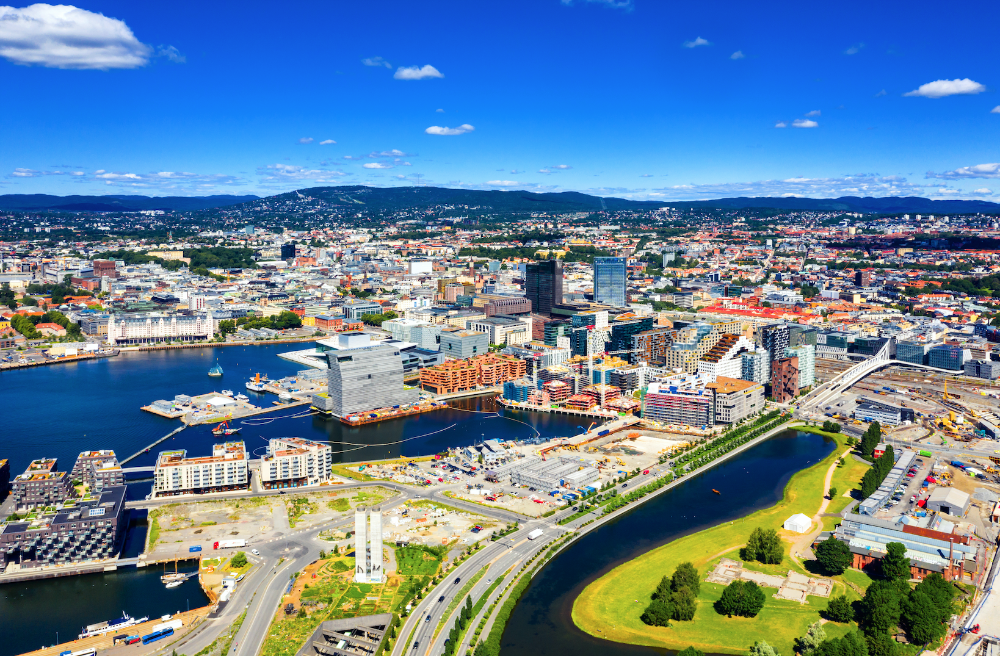 Aerial view of Oslo waterfront skyline with modern buildings, harbor, and curving highway under a bright blue sky