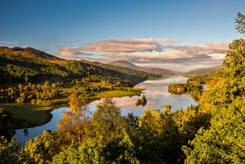 Panoramic view of Queen’s View in Scotland, featuring a serene loch surrounded by forested hills and mountains under a dramatic sky.