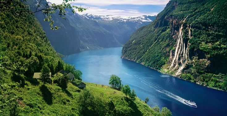 View over Geirangerfjord Norway with steep green mountains, a waterfall, and a cruise boat sailing along deep blue water