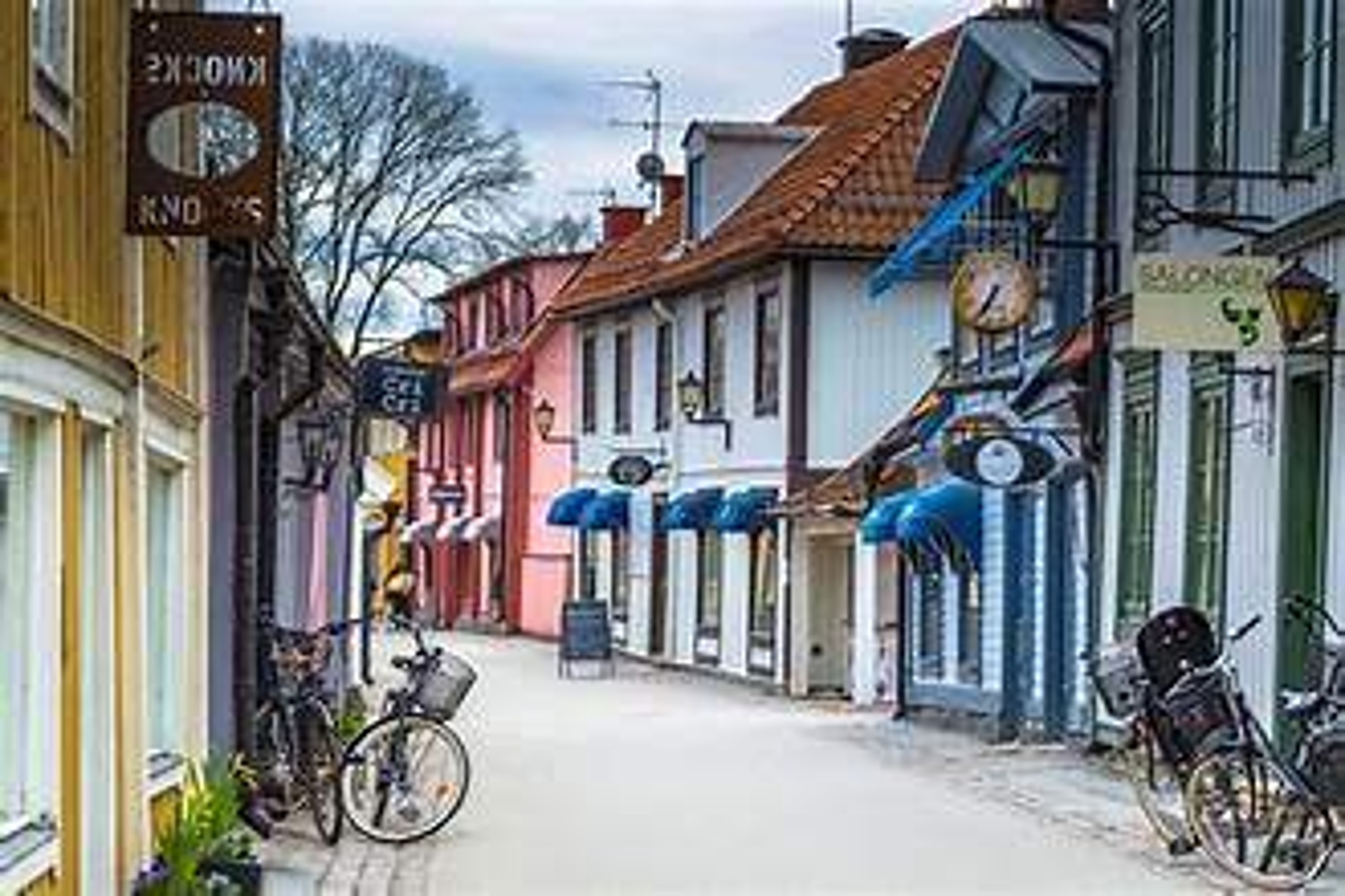 Narrow pedestrian street lined with colorful wooden buildings, shop signs, bicycles, and awnings in a quaint small‑town setting.